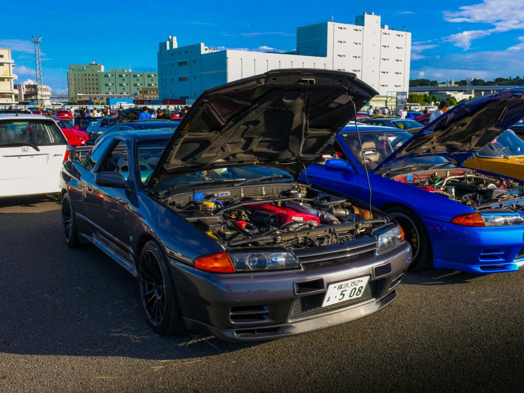 A collection of classic JDM cars with open hoods at an outdoor car meet, showcasing iconic Japanese vehicle culture.